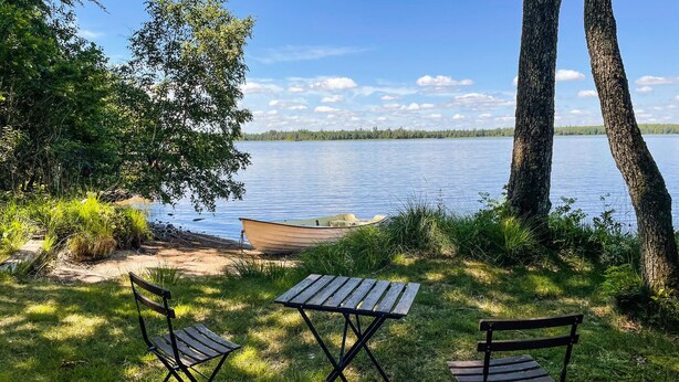 Seeblick mit Holztisch, zwei Stühlen, einem Boot und Bäumen.