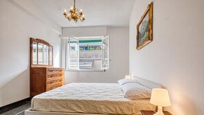 Un dormitorio con cama, cómoda, ventana con vistas despejadas, candelabro y un cuadro colgado en la pared.
