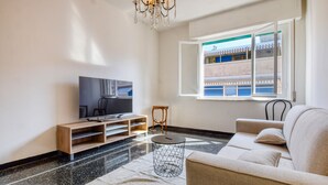 Una sala de estar moderna con televisor de pantalla plana, mueble de entretenimiento de madera, una mesa de centro redonda de metal, un sofá color beige y un ventanal con vista a un edificio azul.