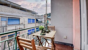 Un balcón con mesa y sillas de madera, una planta en maceta y vistas a edificios con contraventanas azules.