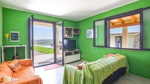 Une pièce aux murs verts, avec une télévision, un canapé et un lit. Une fenêtre offre une vue sur une piscine et un immeuble.