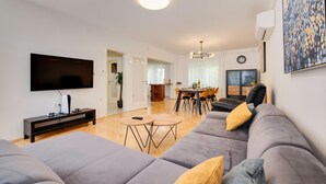 Un salon moderne avec un canapé gris, un téléviseur à écran plat, une table basse et un coin repas comprenant une table et des chaises en bois.