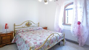 Quarto com colcha florida, mesa de cabeceira com luminária, janela com cortinas e lustre.