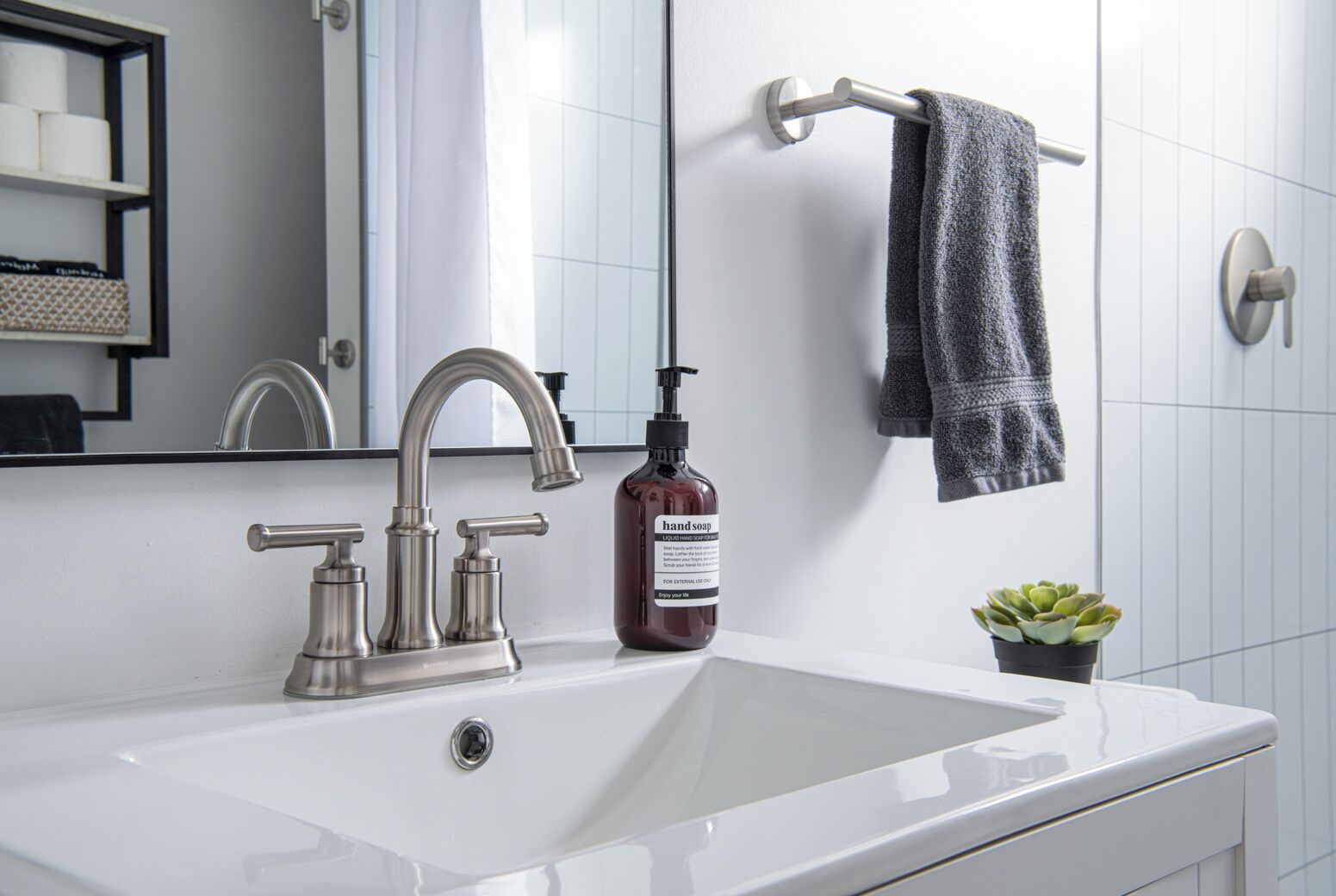 A bathroom with a white sink, a silver faucet, a bottle of hand soap, a towel rack with a towel, and a small potted plant.