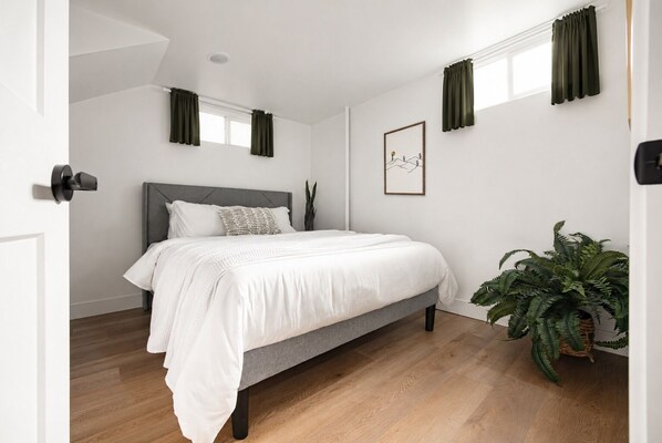 A bedroom with a bed, a potted plant, a framed picture on the wall, and a window with curtains.