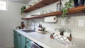 A kitchen with teal cabinets, a stainless steel sink, wooden shelves with kitchenware, and a window providing natural light.