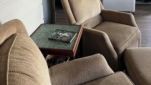 Une cuisine de taille réduite, équipée d’une cuisinière, d’un réfrigérateur et d’une fenêtre avec des rideaux. On y trouve deux fauteuils beiges et une petite table sur laquelle sont posés un livre et des ciseaux.