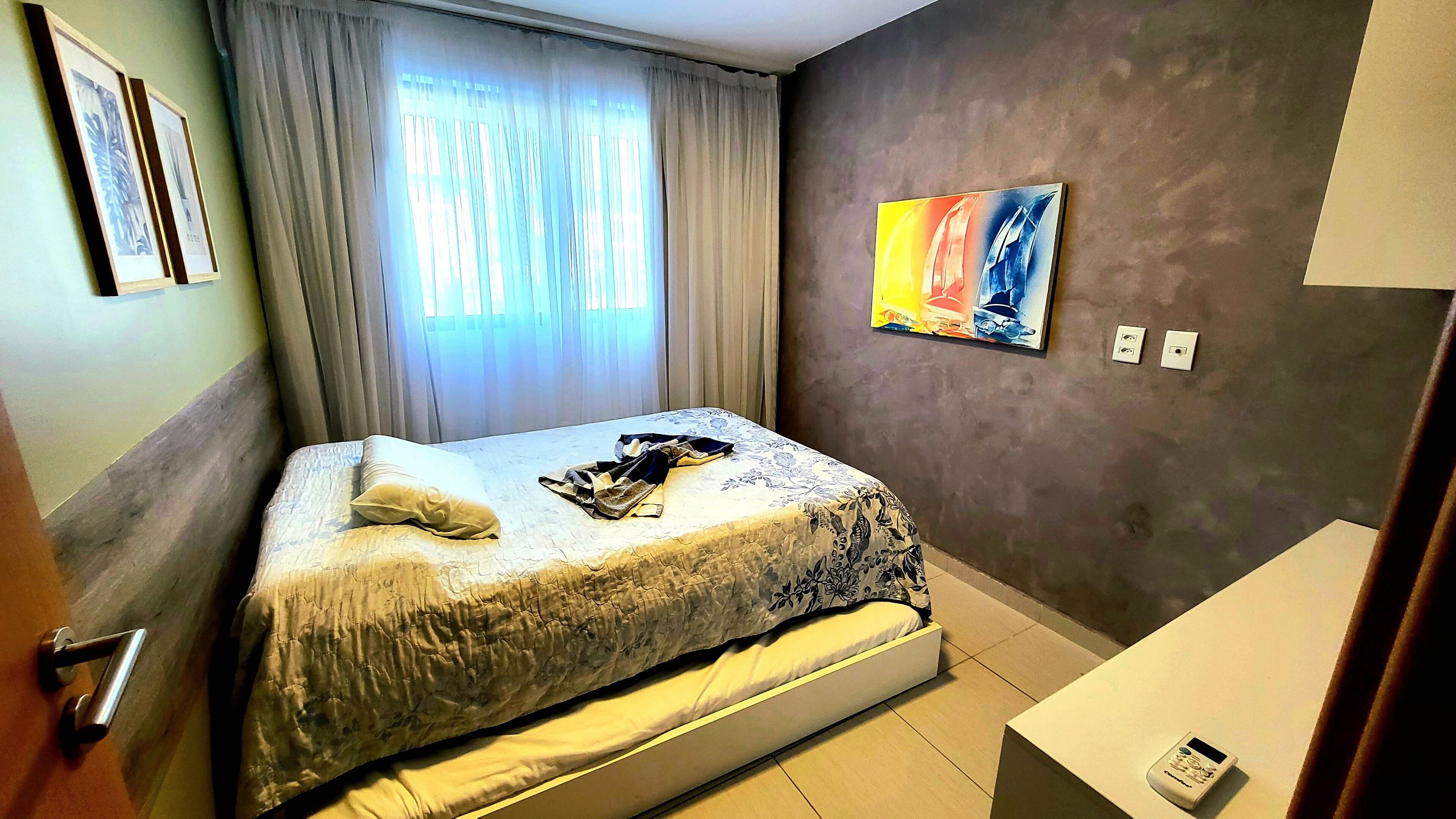 A hotel room with a bed, a window with curtains, a painting on the wall, and a bedside table.