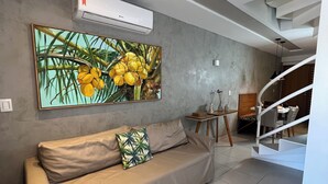 Egy modern nappali, melyben bézs színű kanapé, egy nagyméretű, trópusi virágmotívumot ábrázoló festmény, egy lépcsőház és egy étkező található.