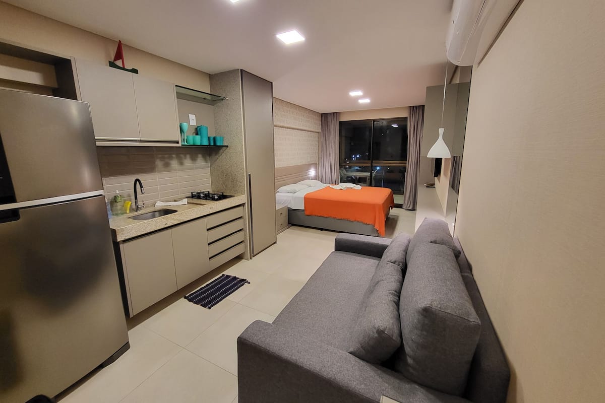 A compact hotel room with a kitchenette, a bed with an orange blanket, a grey sofa, and a large window offering a city view.