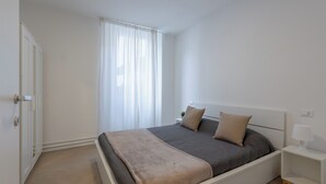 A modern bedroom with a large bed, a bedside table, and a window with sheer curtains.