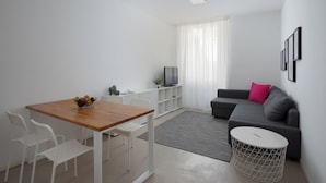 A modern living room with a wooden dining table, white chairs, a grey sofa, and a TV on a white cabinet.