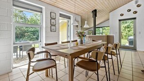 A modern dining area with a long wooden table, multiple chairs, and large windows offering a view of greenery. - Lovely home in Glesborg with sauna (Glesborg)