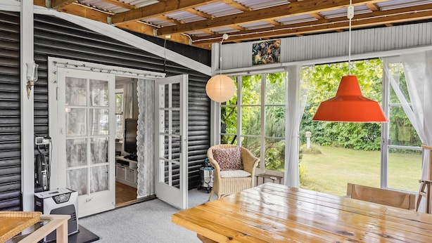 A cozy room with a wooden table, wicker chair, and a large window with a view of greenery. - 2 bedroom cozy home in Faxe (Faxe)