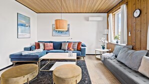 Un salon moderne avec un canapé bleu, un canapé gris, une table ronde et deux poufs ronds. Un luminaire suspendu se trouve au-dessus du canapé bleu, et une horloge est accrochée au mur.