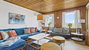 Un salon moderne avec un canapé bleu, un canapé gris, une table basse et deux poufs ronds. Il y a un tableau au mur, une horloge et une fenêtre avec des rideaux.