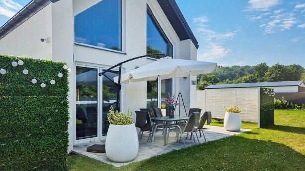 Una casa moderna con un patio que cuenta con una mesa, sillas, un parasol blanco y plantas en macetas.