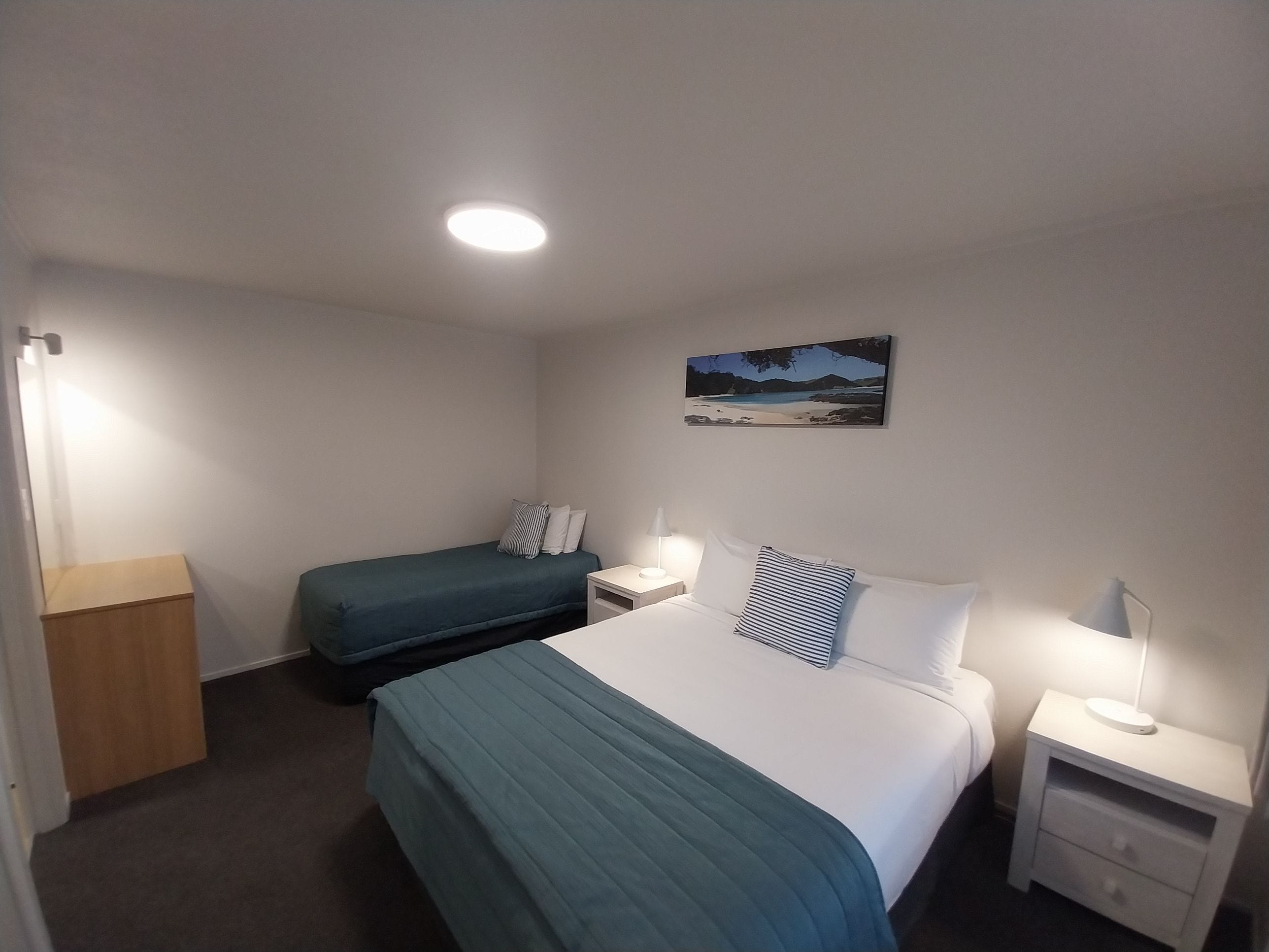 A hotel room with a bed, a smaller bed, a nightstand with a lamp, a wooden cabinet, and a painting on the wall.