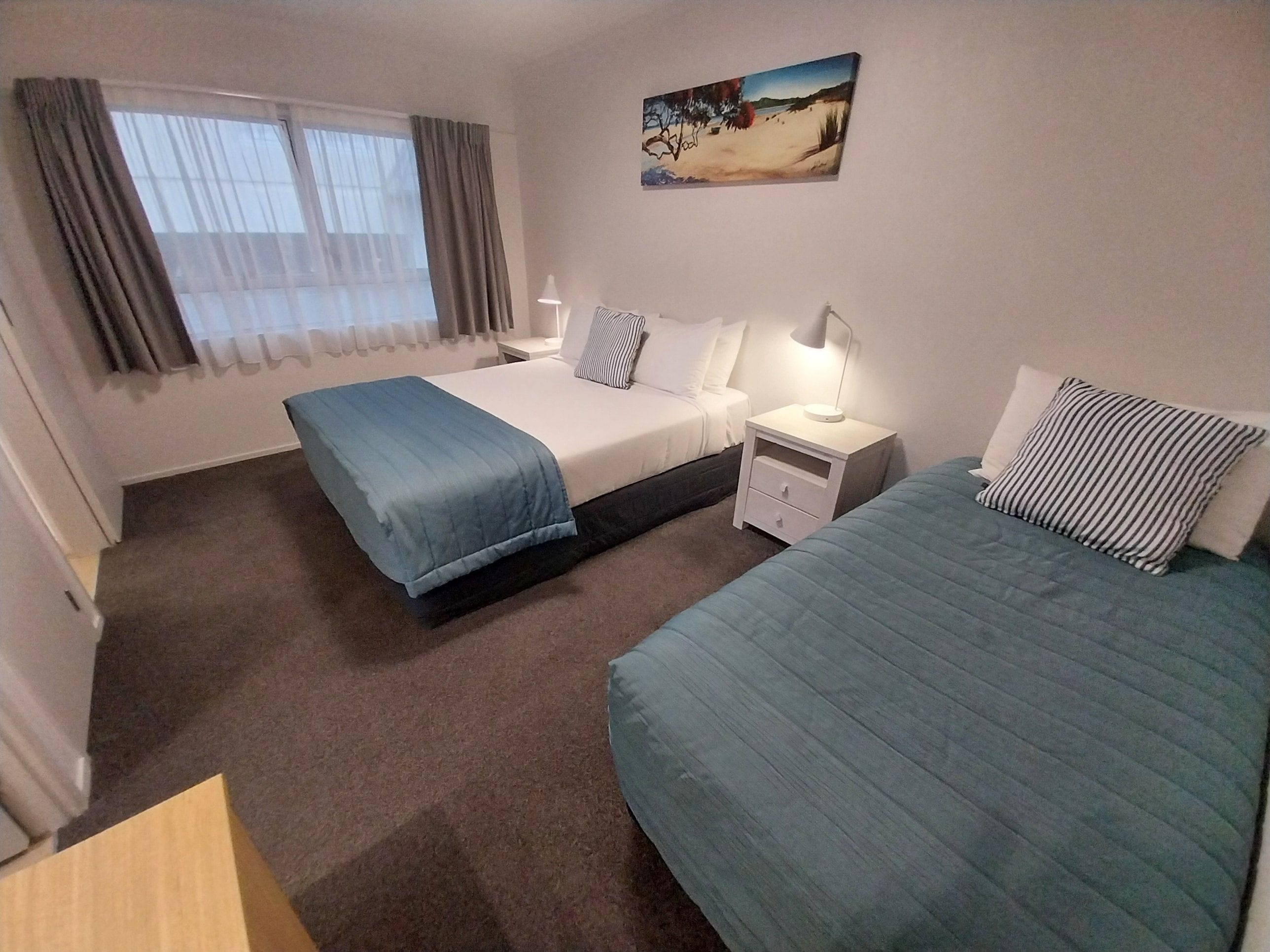 Ein Hotelzimmer mit zwei Betten, einem Nachttisch mit Lampe, einem Bild an der Wand und einem Fenster mit Vorhängen.