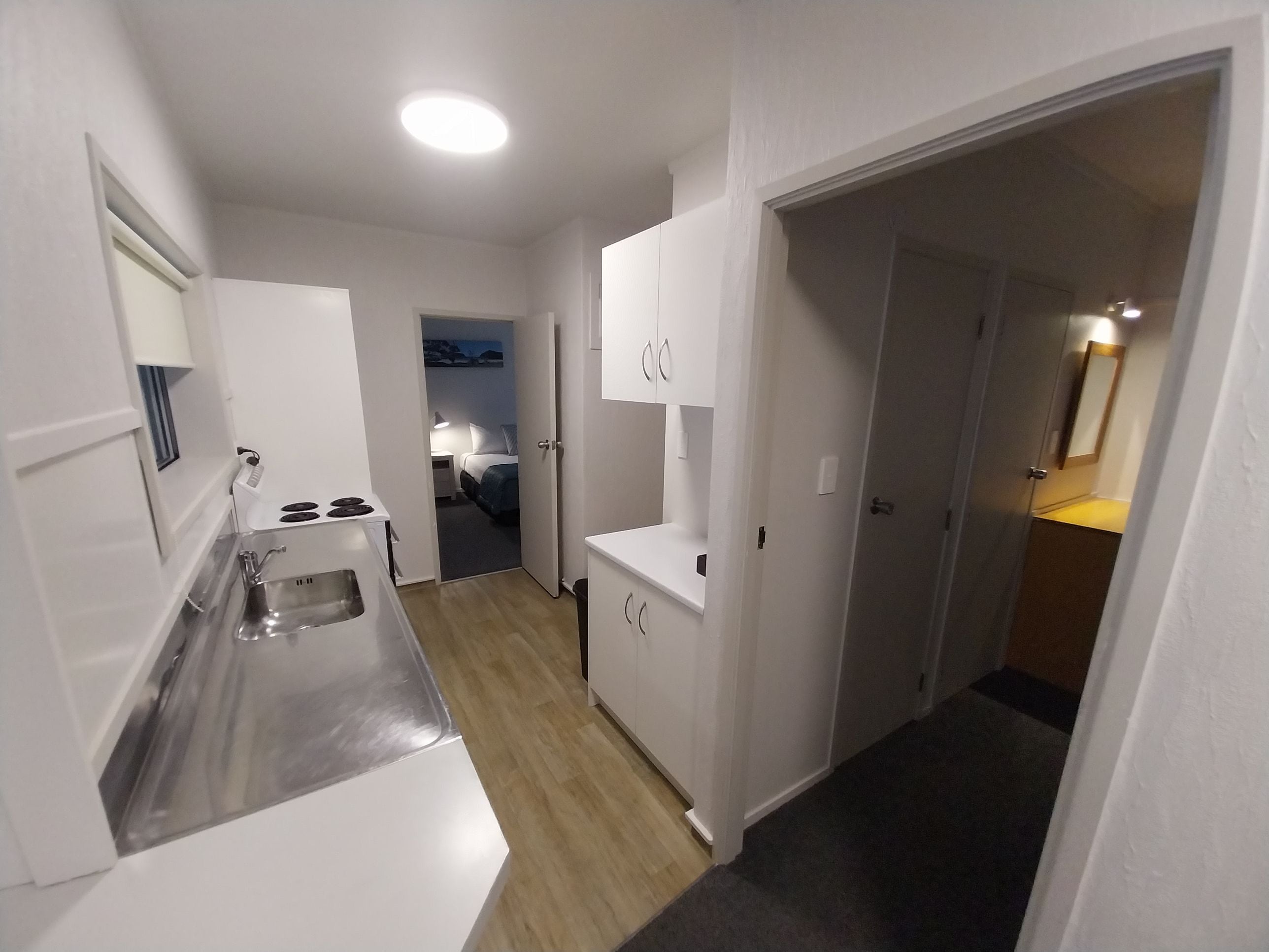 Een compacte keuken met een spoelbak, fornuis en kastjes, die uitkomt op een slaapkamer met een bed en nachtkastje.