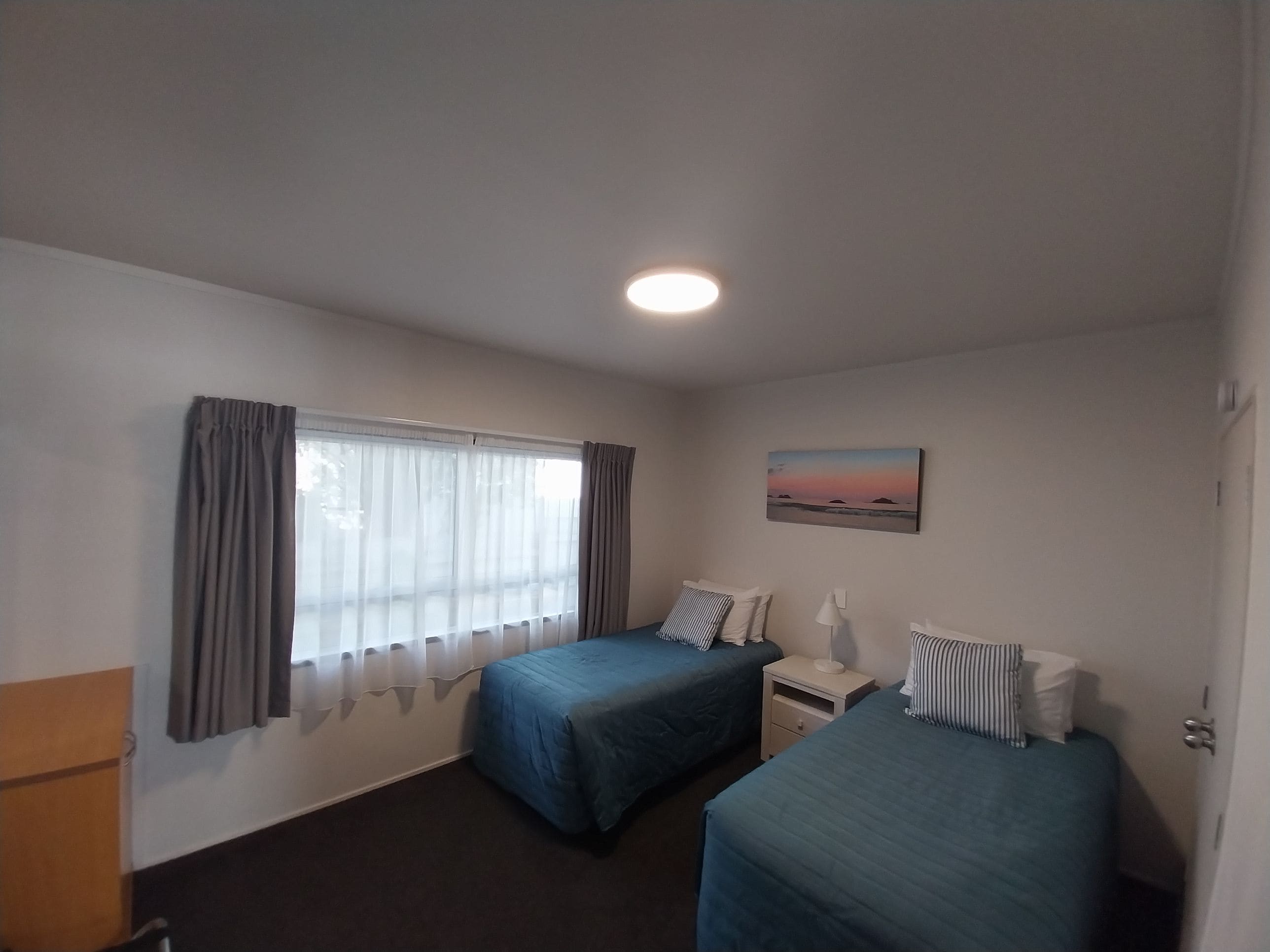 A room with two beds, a window with curtains, a bedside table with a lamp, and a painting on the wall.