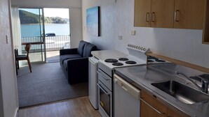 A compact kitchen with a stove, sink, and wooden cabinets. There is a dining area with a table and chairs, and a view of a body of water through a glass door. - Bayview Motel (Paihia)
