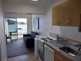 Una cocina compacta con estufa, pileta y muebles de madera. Hay un comedor con mesa y sillas, y vista a una masa de agua a través de un vidrio.