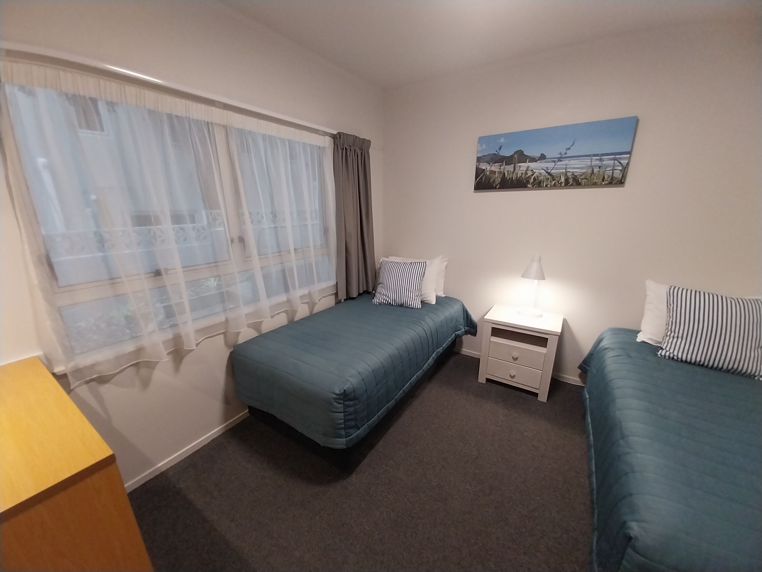 Un dormitorio con dos camas individuales, una mesita de noche con lámpara, una ventana con cortinas traslúcidas y un cuadro en la pared.
