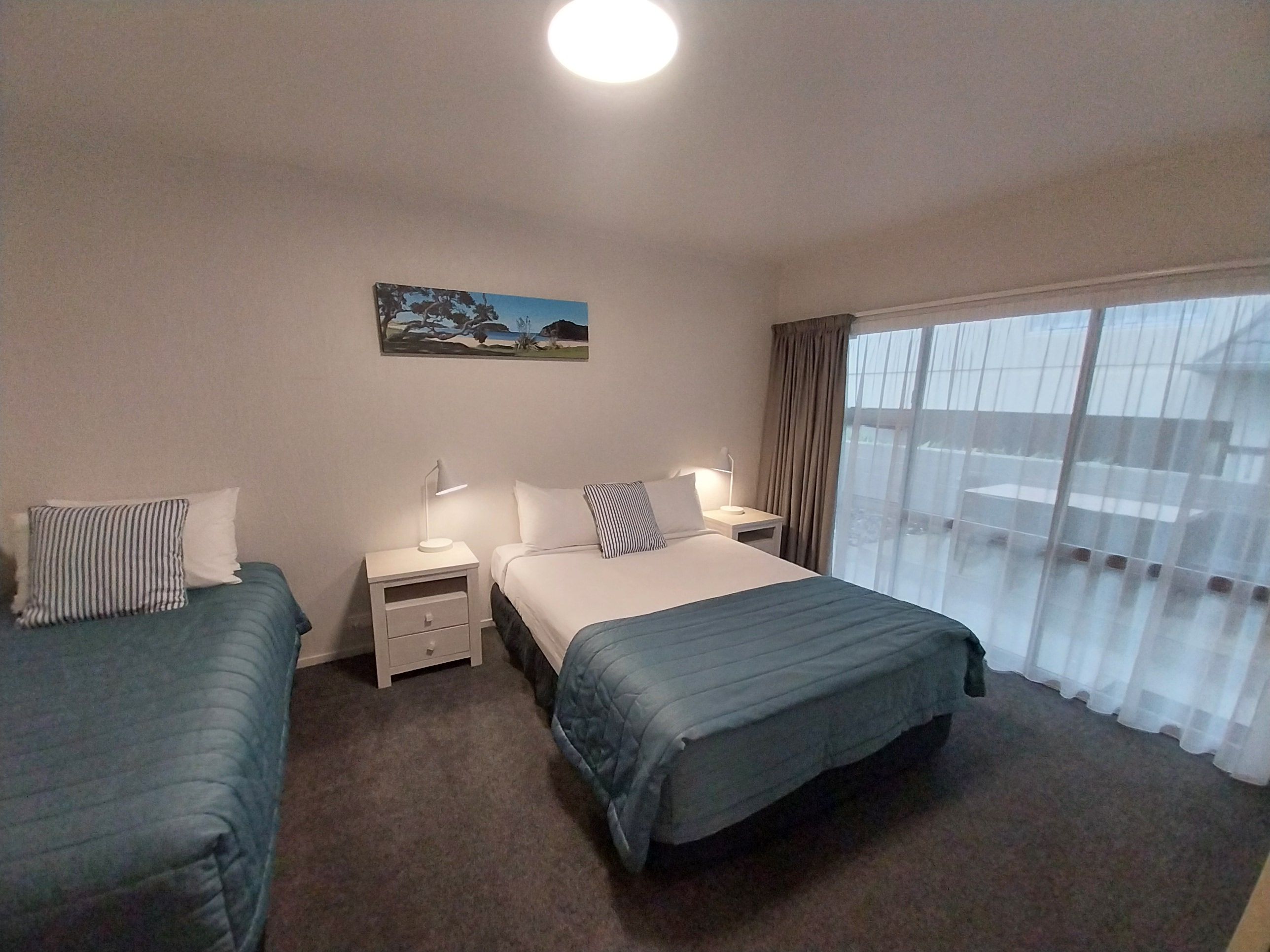 A hotel room with two single beds, a nightstand with a lamp, a painting on the wall, and a large window with curtains.