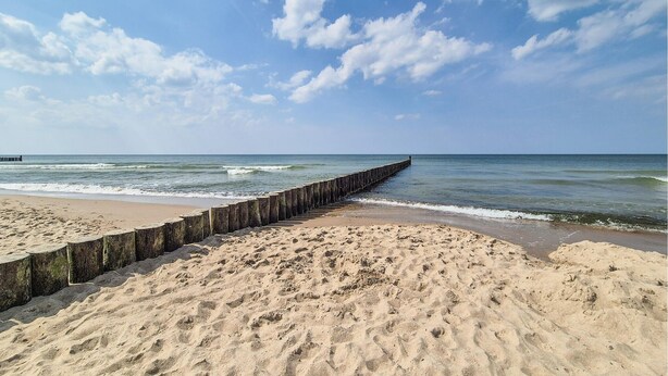 A beach with a wooden jetty extending into the ocean, sandy terrain, and a clear sky with scattered clouds.