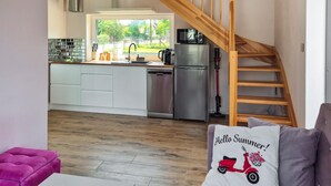 A modern kitchen with a wooden staircase, a dining area with a purple sofa, and a view of greenery outside.