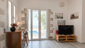 Une pièce avec un bureau en bois, un téléviseur posé sur un meuble en bois, une porte donnant sur un balcon avec vue sur la mer, et une petite plante accrochée au mur.