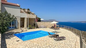 Un espace piscine doté d’un sol en pierre, de chaises longues et de parasols, offrant une vue sur la côte.