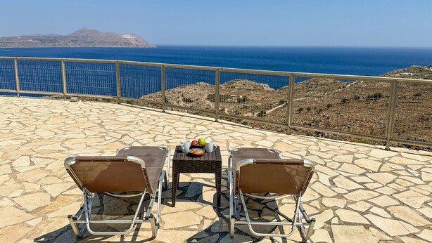 Une terrasse avec deux chaises et une table à structure métallique, offrant une vue dégagée sur un paysage côtier, la mer et les collines lointaines.