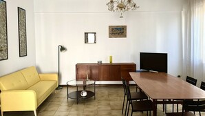 Olohuoneessa on sohva, pyöreä sohvapöytä, televisio, ruokapöytä tuoleineen, kattokruunu ja seinällä on kehystettyjä tauluja.