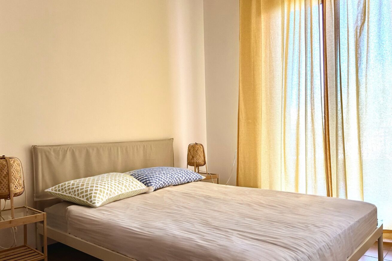 A bed with a headboard, two pillows, a bedside table with a lamp, and a window with curtains.