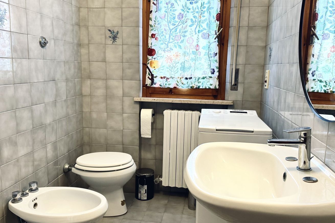 A bathroom with a toilet, bidet, sink, and a window with floral curtains.