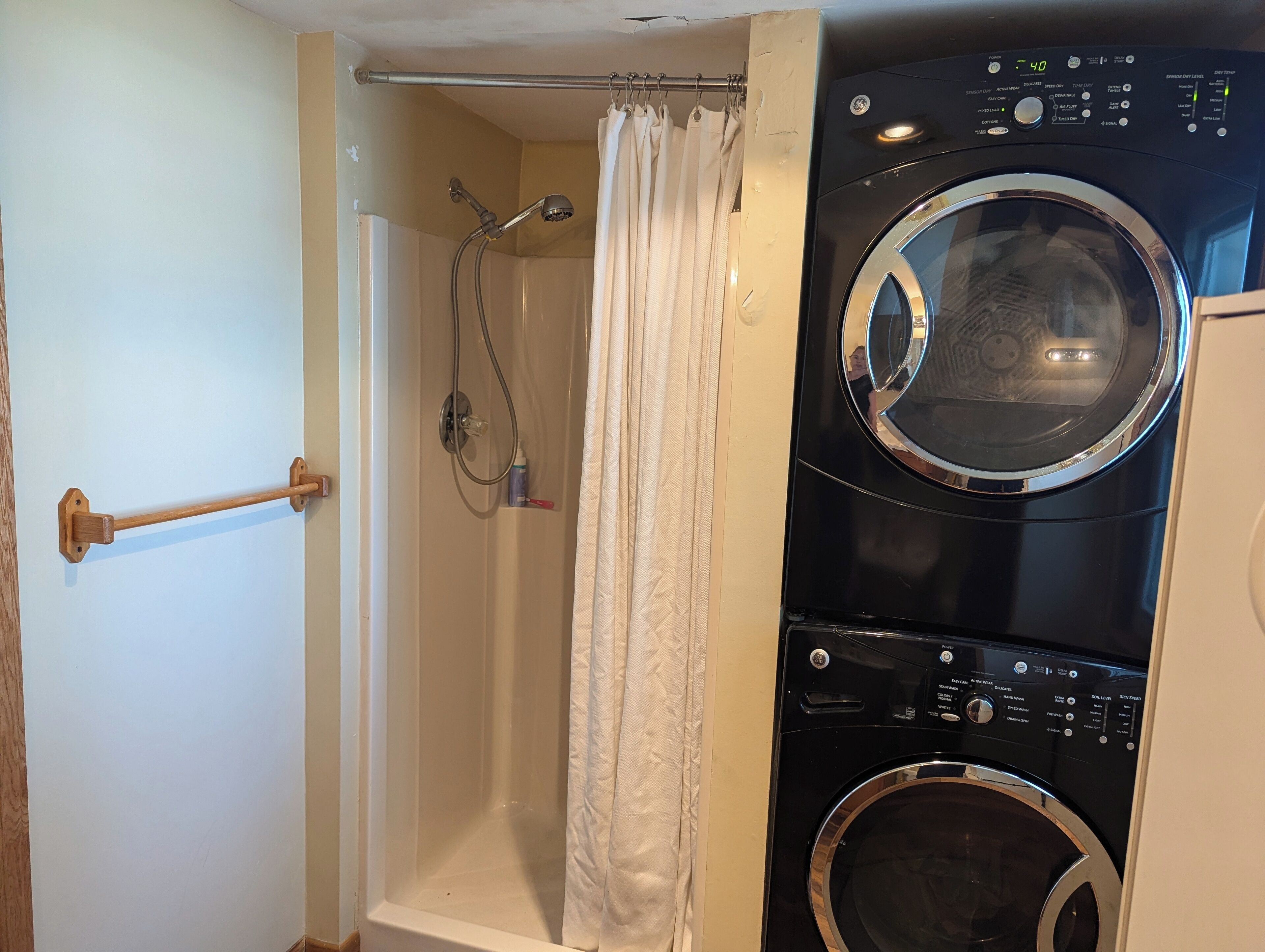 A compact laundry room with a front-loading washer and dryer, a shower with a curtain, and a towel rack.