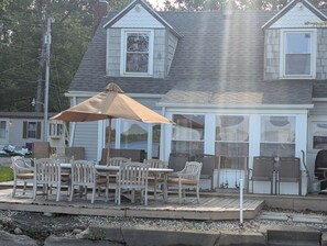Un espace extérieur abrité, aménagé pour les repas, avec une table et des chaises, un grand parasol et une vue sur une maison dotée d’un garage et d’un porche couvert.