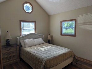 Une chambre à coucher comprenant un lit, deux tables de chevet, une commode, une fenêtre avec des stores et une fenêtre décorative à six pans au-dessus du lit.