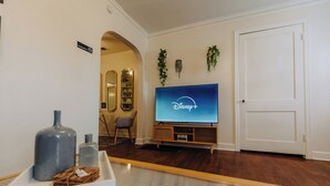 A living room with a flat-screen TV displaying the Disney+ logo, a wooden entertainment unit, a white door, and a small plant on the wall. - Historic Revival King Bed Liberty Bowl Memphis 895 (Memphis)