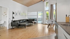 A modern living room with a leather sofa, a dining area with two chairs, a kitchen with a stainless steel sink, and large windows allowing natural light. - Awesome home in Væggerløse with sauna (Væggerløse)