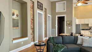 Una sala de estar con un sofá gris oscuro, una mesita redonda y una manta verde. Se aprecia una pared de ladrillo decorativa y un ventilador de techo.