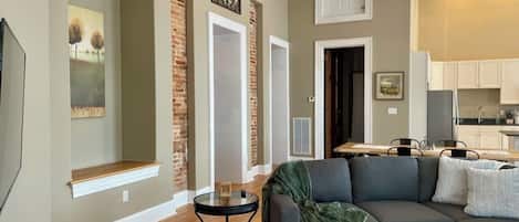 A living room with a dark gray sofa, a small round table, and a green throw blanket. There is a brick accent wall and a ceiling fan.