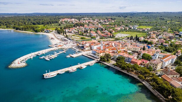 Vista aérea de un pueblo costero con un puerto deportivo, edificios con techos de tejas rojas y un mar azul cristalino.