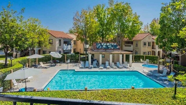 Uma área de piscina com espreguiçadeiras e guarda-sóis, cercada por edifícios com varandas.