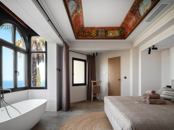 A bedroom with a large window, a bathtub, a bed with towels, and a wooden door.