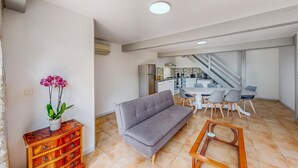 Ein Wohnzimmer mit grauem Sofa, Holztisch und einer Kommode aus Holz mit Topfpflanze. Eine Treppe führt zu einer Küche und einem Essbereich mit gefliestem Rücksprung und modernen Geräten.