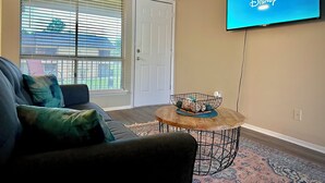 A living room with a TV mounted on the wall displaying the Disney+ logo, a sofa with cushions, a coffee table with decorative items, and a patterned rug. - Worker friendly, cozy, great location, quiet, full kitchen, WiFi,  (Sherman)