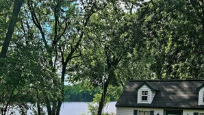 A small white house with a green door and windows, surrounded by a white fence and trees. There are three kayaks in blue, orange, and gray colors on the grass.